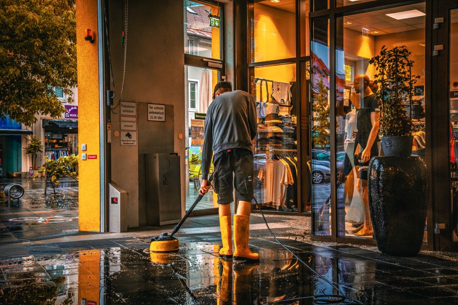 A person wearing yellow rubber boots is engaged in surface cleaning outside a retail shop. They are using a scrubber or floor cleaner on a wet, reflective sidewalk, which appears to be cleaned recentl
