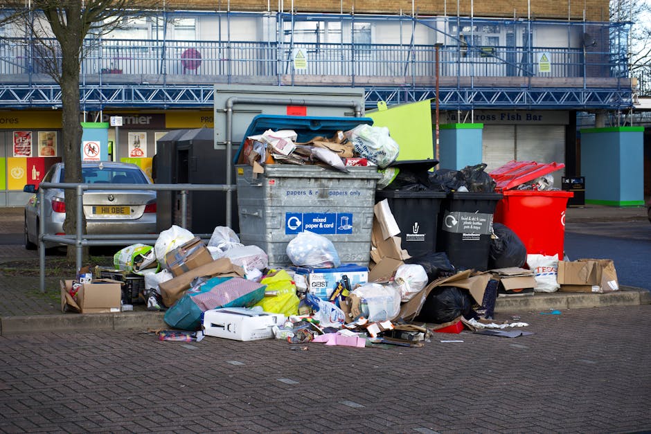 A waste collection worker wearing a high-visibility yellow vest and dark clothing is operating a refuse vehicle on a city street during dusk or early evening. The vehicle has a large, open rear compar