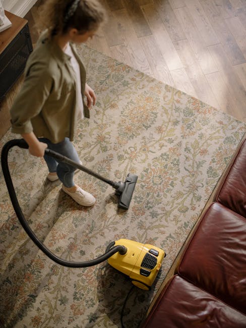 A woman is engaged in deep surface cleaning on a patterned, beige and green area rug in a living room with wooden flooring. She is using a yellow and black vacuum cleaner, holding the hose attachment 